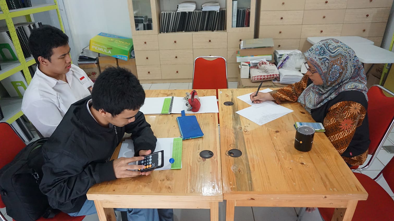Two blind students sit on the left side of a table, each with Braille paper and Braille writing slates in front of them. One student is using a mobile phone calculator. On the right side of the table, a teacher is writing on a piece of paper with a pencil.
