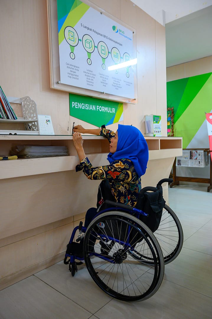 A woman in a wheelchair in front of a table with forms. The table is too high. The woman cannot reach the forms or fill them out.